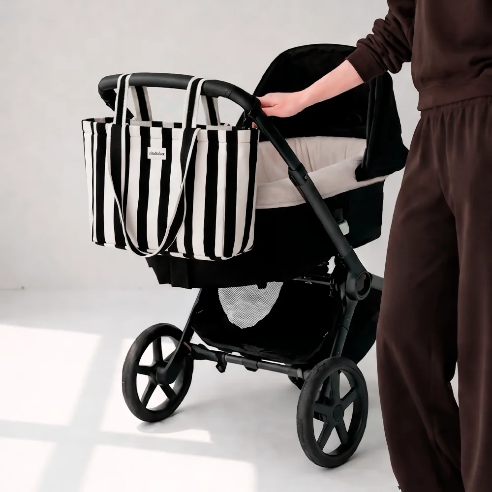 Bolsa de carro de bebé Mum Rayas Negras versátil y elegante, cierre con cremallera y asas múltiples. El bolso de carro Mum rayas negras de rita&deer lleva cierre con cremallera y una correa que puedes colgarla con su doble asa con automáticos, lo que lo convierte en el bolso de carrito perfecto para llevar tus pertenencias de manera segura. Además, sus dos asas largas te permite llevarlo cómodamente al hombro como un tote bag durante tus paseos con tu bebé. Gracias a su espacio interior y sus dos compartimentos, nuestro bolso panera Mum ofrece la capacidad suficiente para llevar todo lo que necesita tu bebé en un solo bolso: pañales, biberones, ropa de repuesto, una mudita y otros accesorios esenciales del bebé